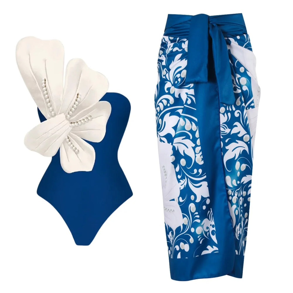 Blue one-piece swimsuit with white floral embellishment and pearl details, paired with a blue and white floral wrap skirt.