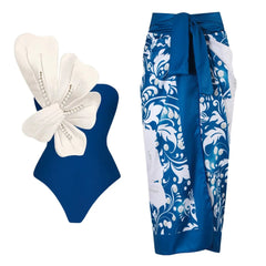 Blue one-piece swimsuit with white floral embellishment and pearl details, paired with a blue and white floral wrap skirt.