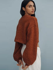 Woman wearing a brown knitted sweater and white pants against a gray background