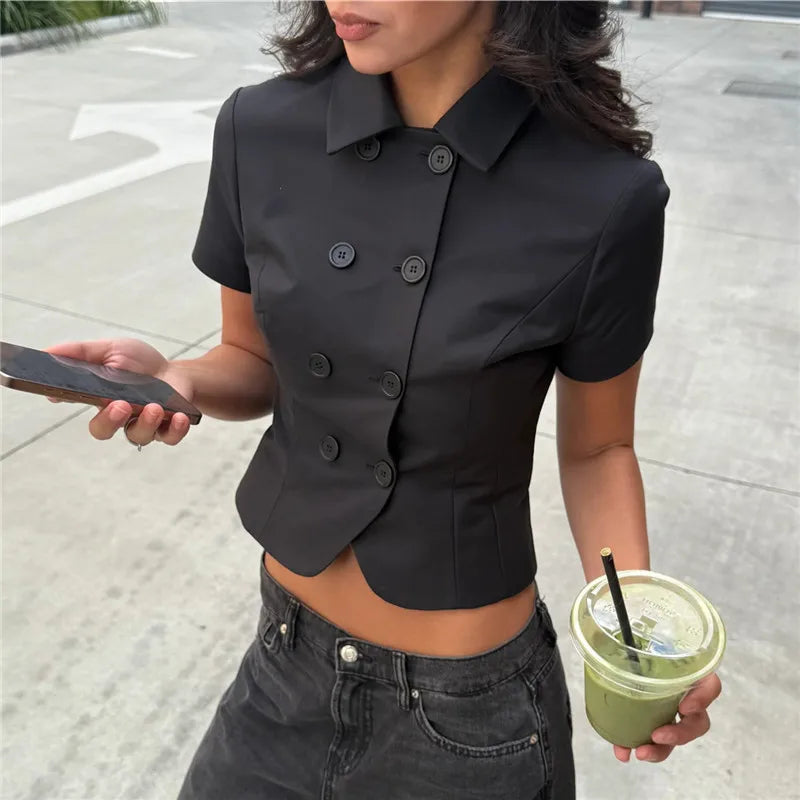 A person wearing a black short-sleeve top holding a drink and phone on a street.