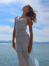 Woman in a light dress with beaded details standing by a body of water under a blue sky.