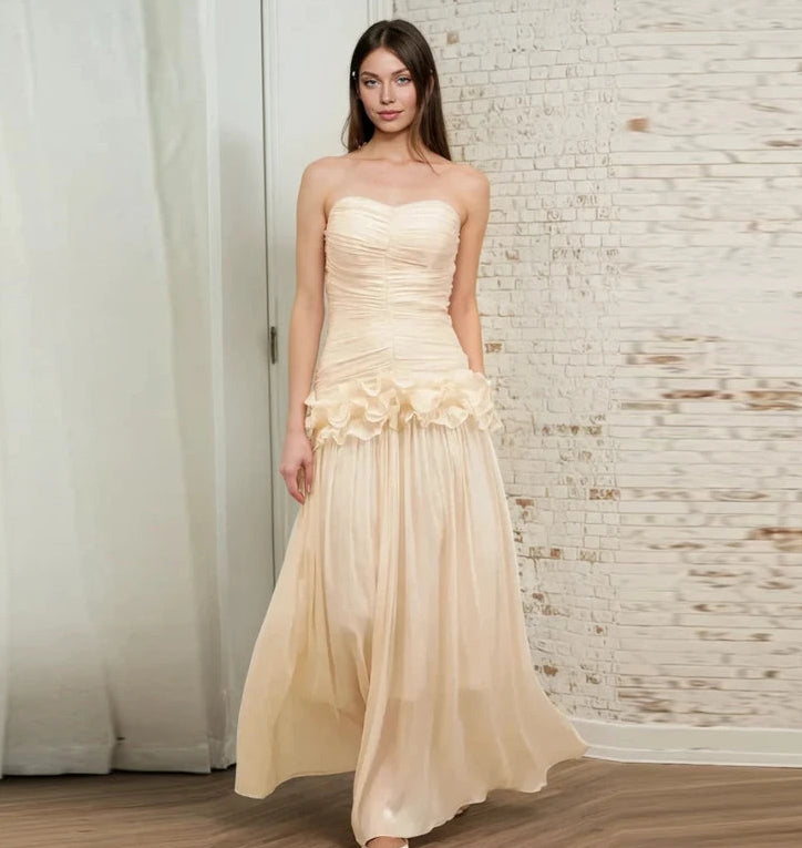 Woman wearing a strapless beige evening gown with floral details against a textured wall.
