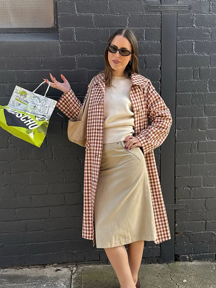 Woman in a checkered coat and beige skirt holding a green and white bag against a black brick wall.