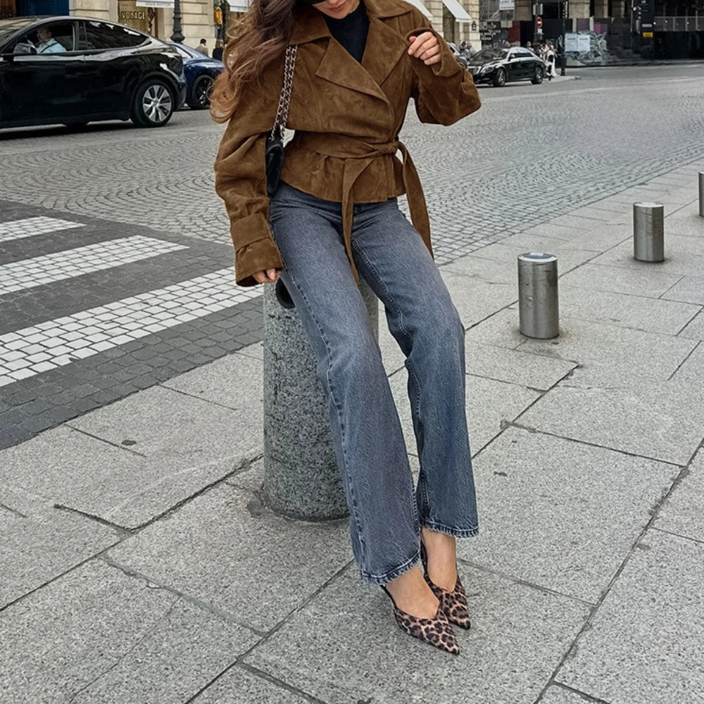 Person wearing a brown jacket and blue jeans standing on a city street.