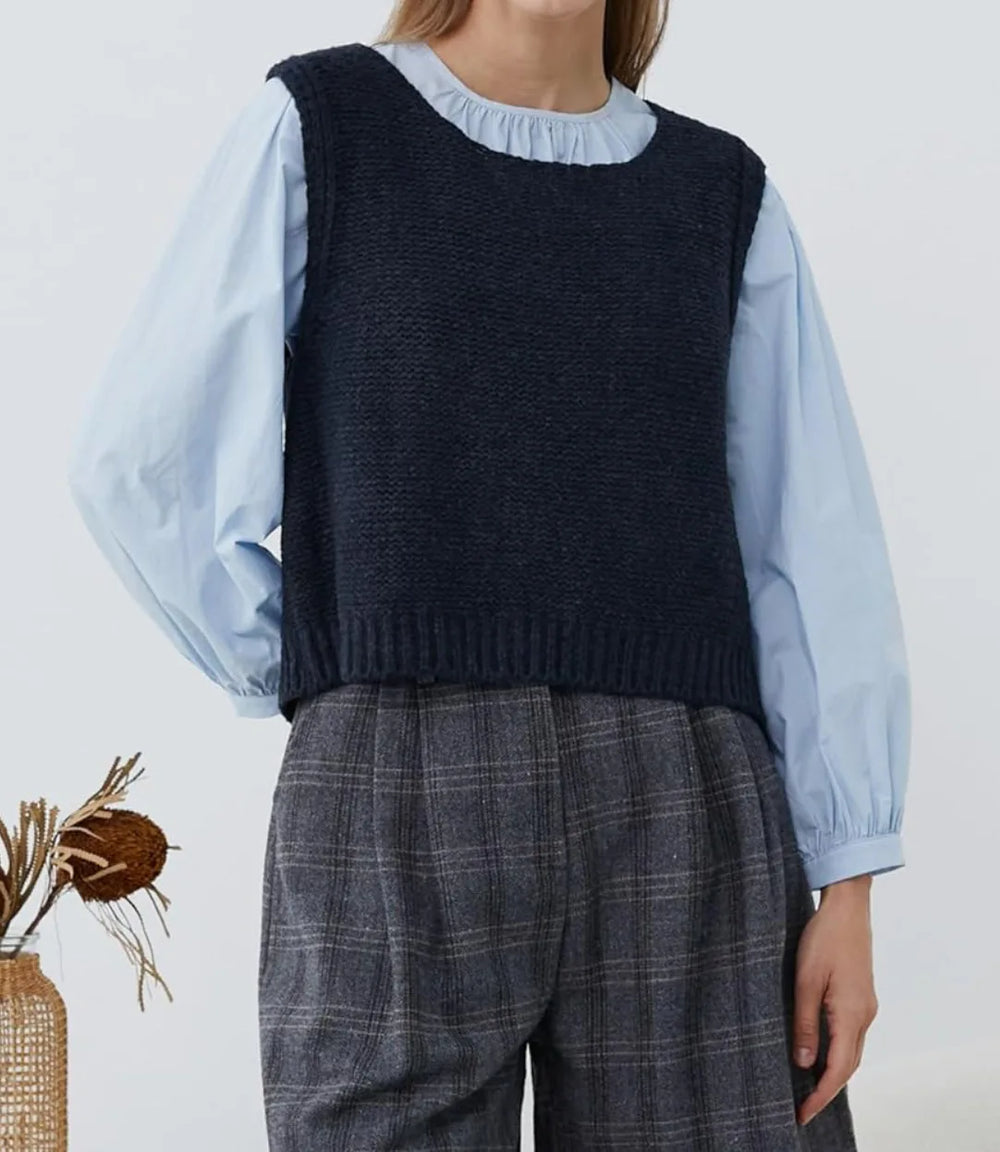 Person wearing a navy knitted vest over a light blue shirt with plaid pants on a plain background