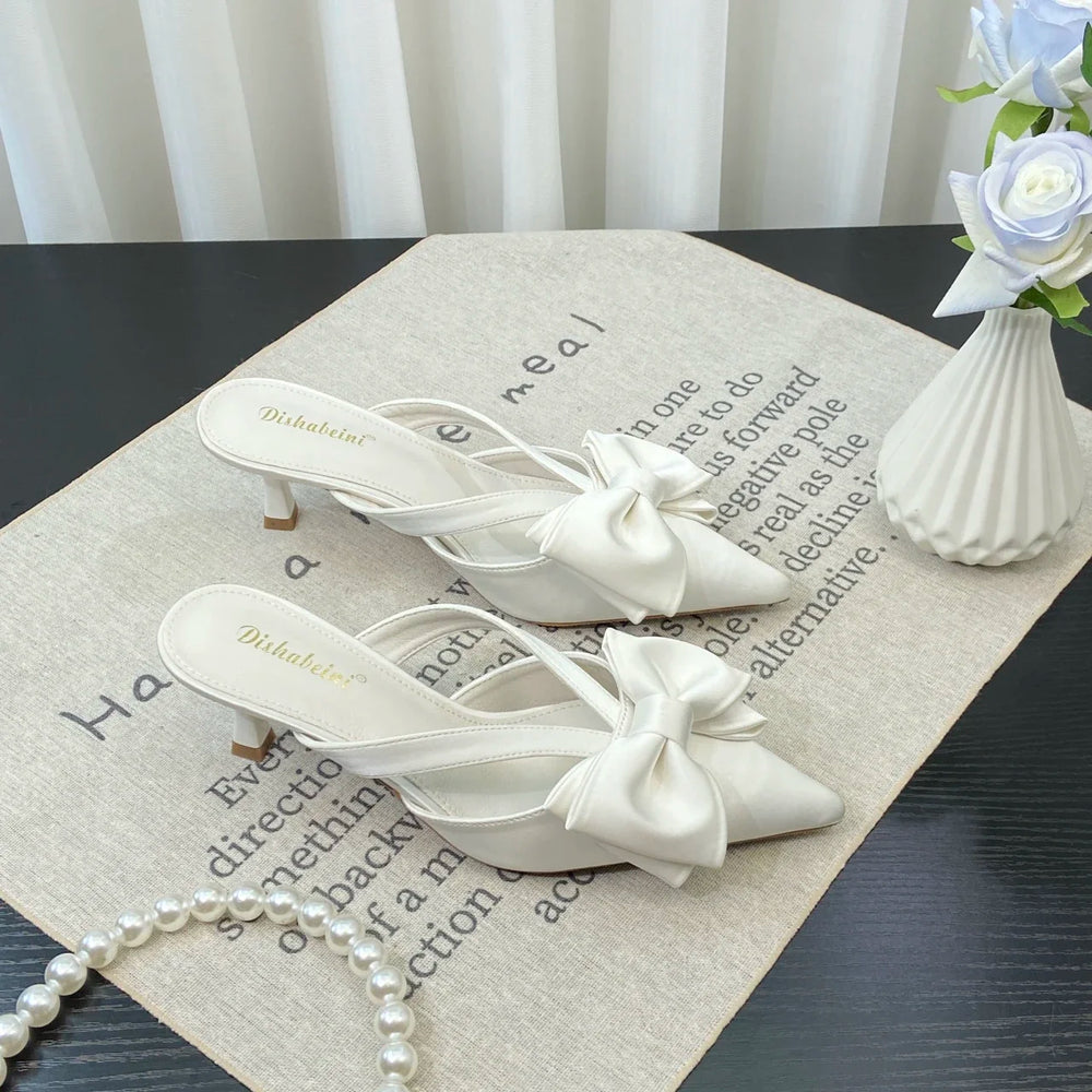 White high-heeled shoes with bows on a textured surface with a vase of flowers in the background.