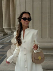 Woman in a white coat and sunglasses holding a beige handbag in front of classical architecture.