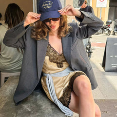 Woman sitting on a bench wearing a blue cap, gray blazer, and beige dress with lace details.