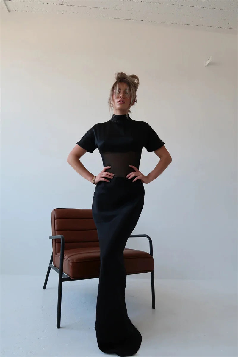 Person wearing a black dress standing in front of a white wall with a brown chair.