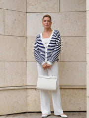 Woman wearing a striped cardigan, white shirt, and pants, holding a white handbag against a beige wall.