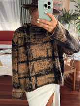 Person wearing a plaid sweater taking a mirror selfie in a room with furniture and plants.