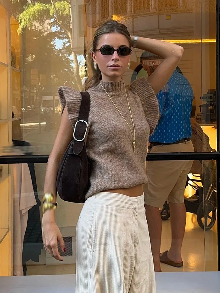 Woman wearing a brown knit top and white pants standing in front of a store window.