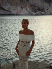 Woman in a white lace dress standing by a body of water with mountains in the background
