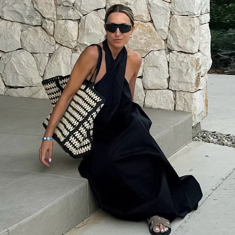 Woman in a black dress and sunglasses holding a patterned bag against a stone wall.