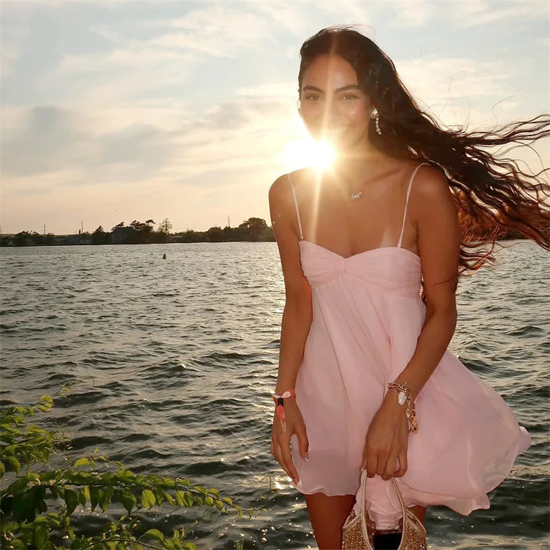 Woman in a pink dress standing by a body of water with the sun shining through her hair.