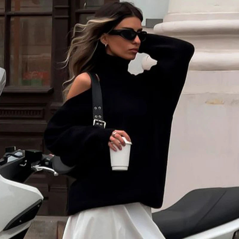 Woman in a black oversized sweater and sunglasses holding a coffee cup, standing next to a motorcycle.