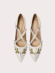 Pair of white pointed-toe shoes with floral embroidery and criss-cross straps on a light gray background.