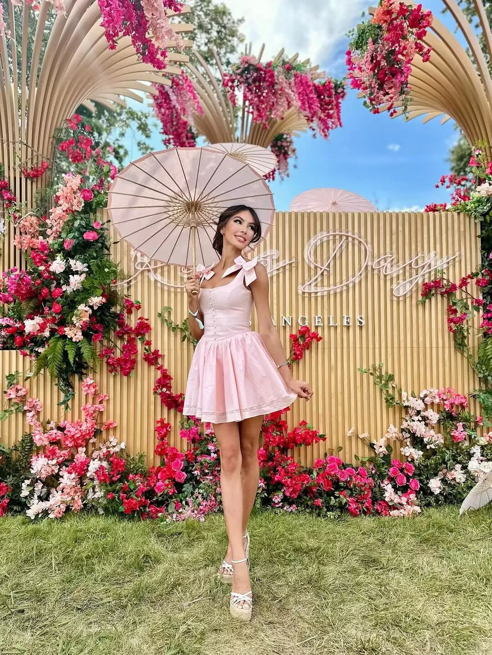 Woman in a pink dress holding an umbrella under a floral arch with 'Daisy' branding.