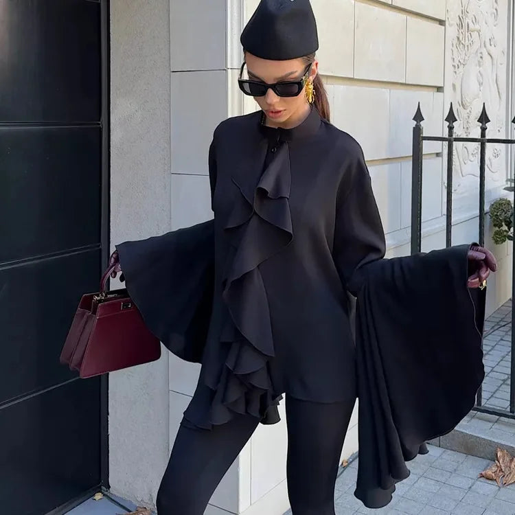 Person wearing a black outfit with ruffled details, holding a maroon handbag, standing against a building.