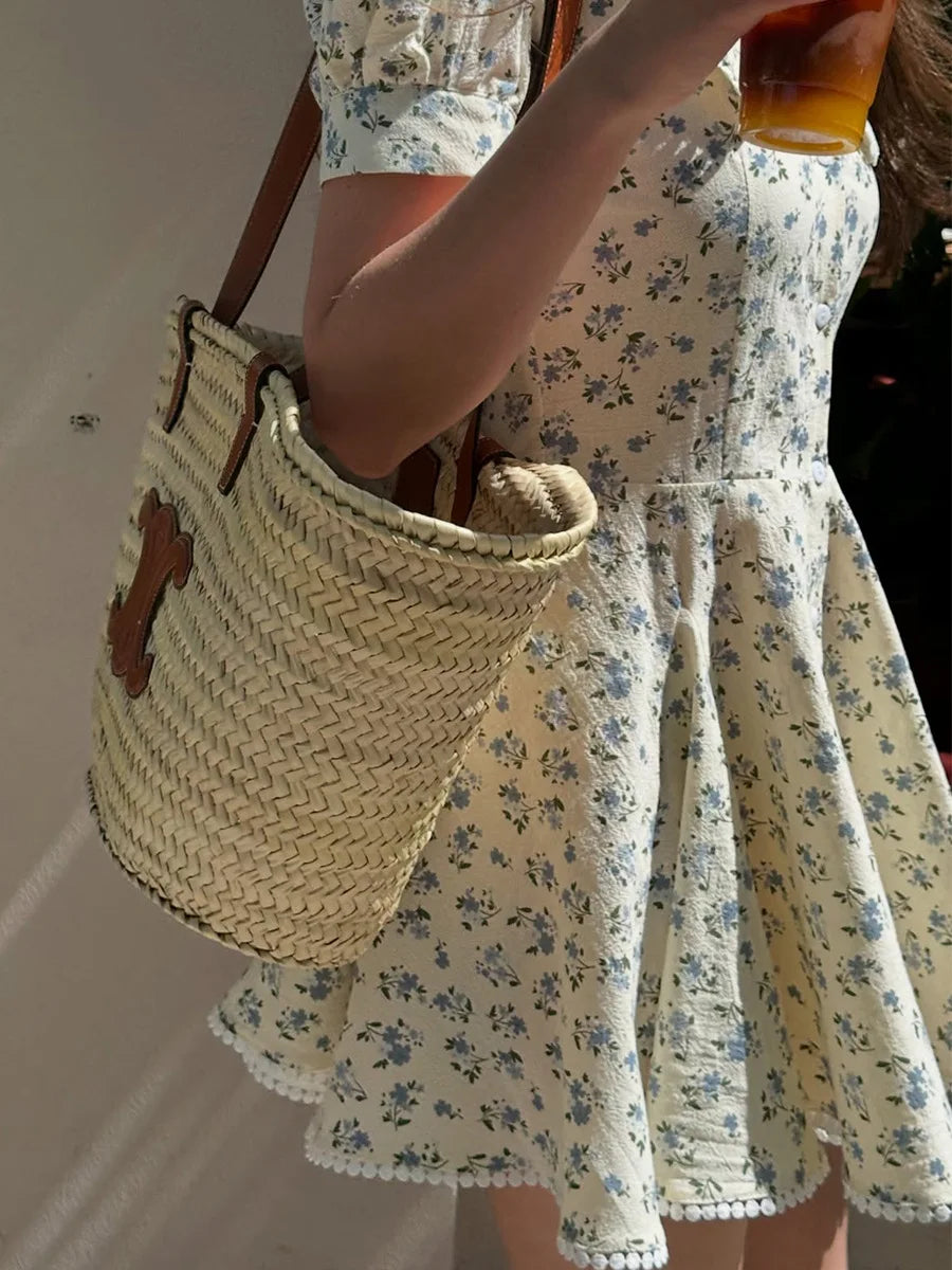 Person wearing a floral dress holding a woven bag and a drink.