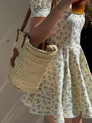 Person wearing a floral dress holding a woven bag and a drink.