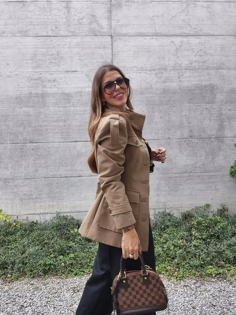 Woman in a beige coat and sunglasses holding a brown handbag against a concrete wall.