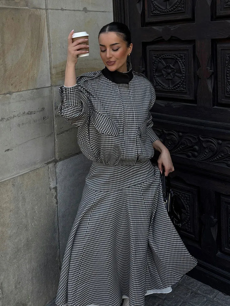 Woman in a checkered dress holding a coffee cup against a stone wall.