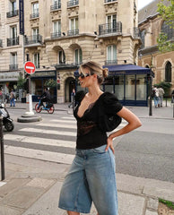 Woman in black top and blue jeans standing on a city street with buildings and pedestrians in the background.