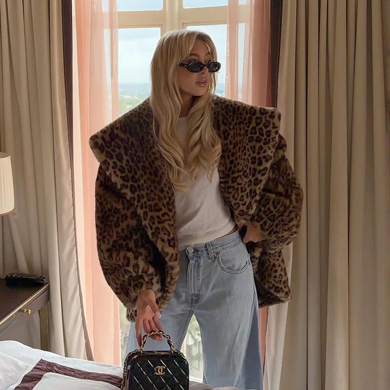 Woman in leopard print coat and sunglasses holding a black handbag in a room with large windows.