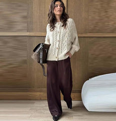 Woman wearing a cream knitted cardigan and dark pants, holding a brown leather bag against a wooden paneled wall.