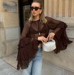 Woman in a brown ruffled blouse and blue jeans holding a white clutch against a stone wall.