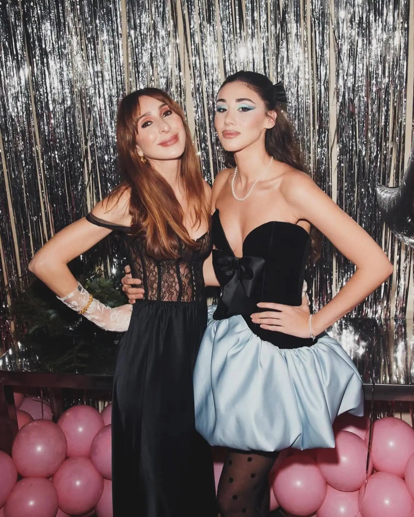 Two women posing together in front of a sparkly curtain with pink balloons.