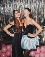 Two women posing together in front of a sparkly curtain with pink balloons.