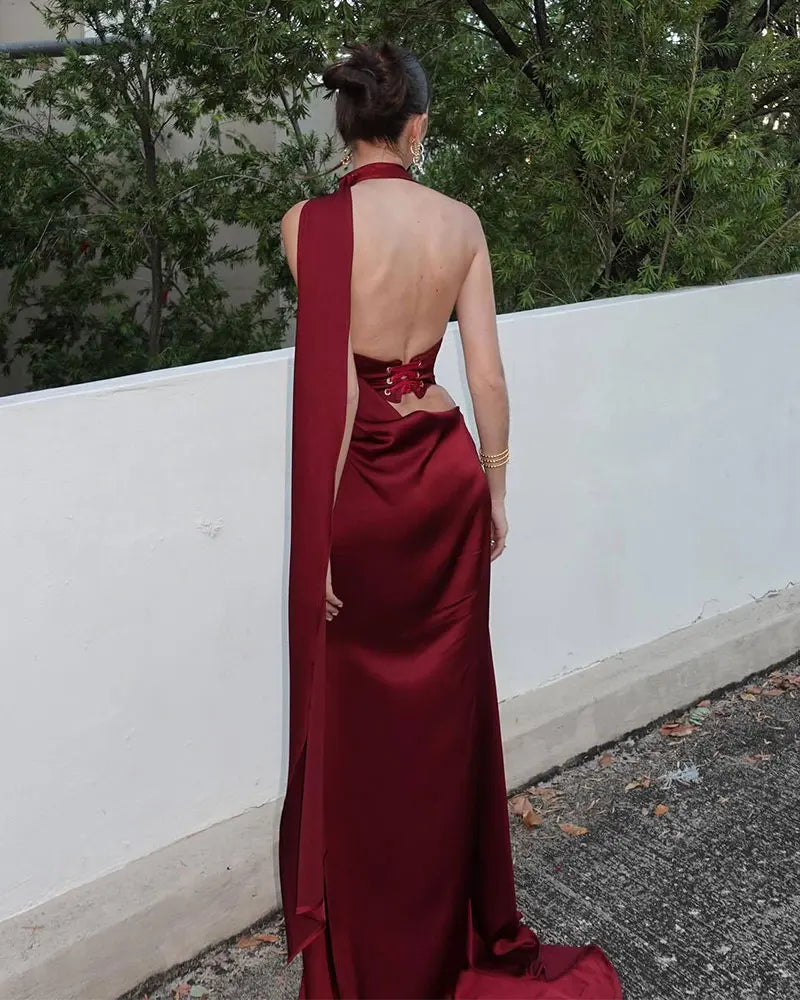Person wearing a burgundy evening gown with a slit, standing against a white wall with greenery in the background.