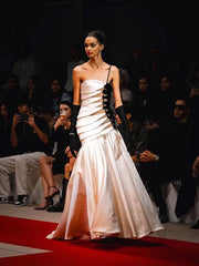 Model wearing a white evening gown on a red carpet with an audience in the background