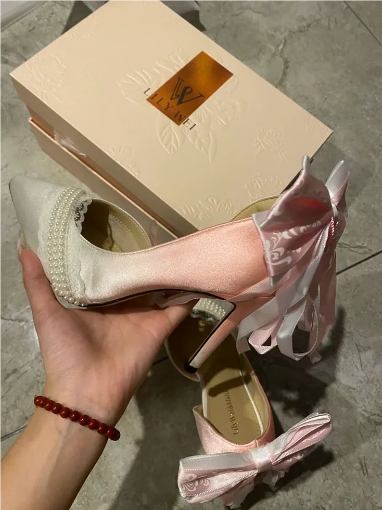 Pink high-heeled shoes with a bow held by a hand, in front of a beige shoe box.