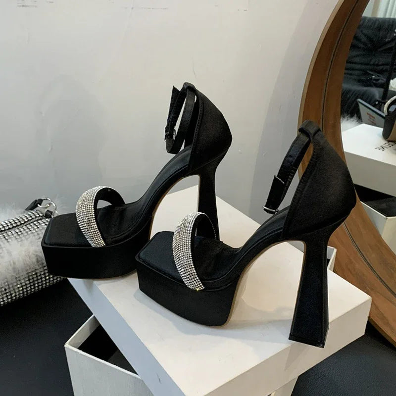 Black high-heeled shoes with rhinestone straps on a white platform, with a mirror and handbag in the background.
