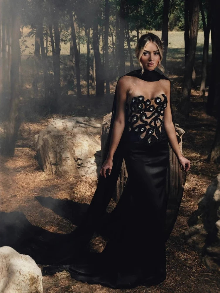 Woman in a black gown with intricate design standing in a forest.