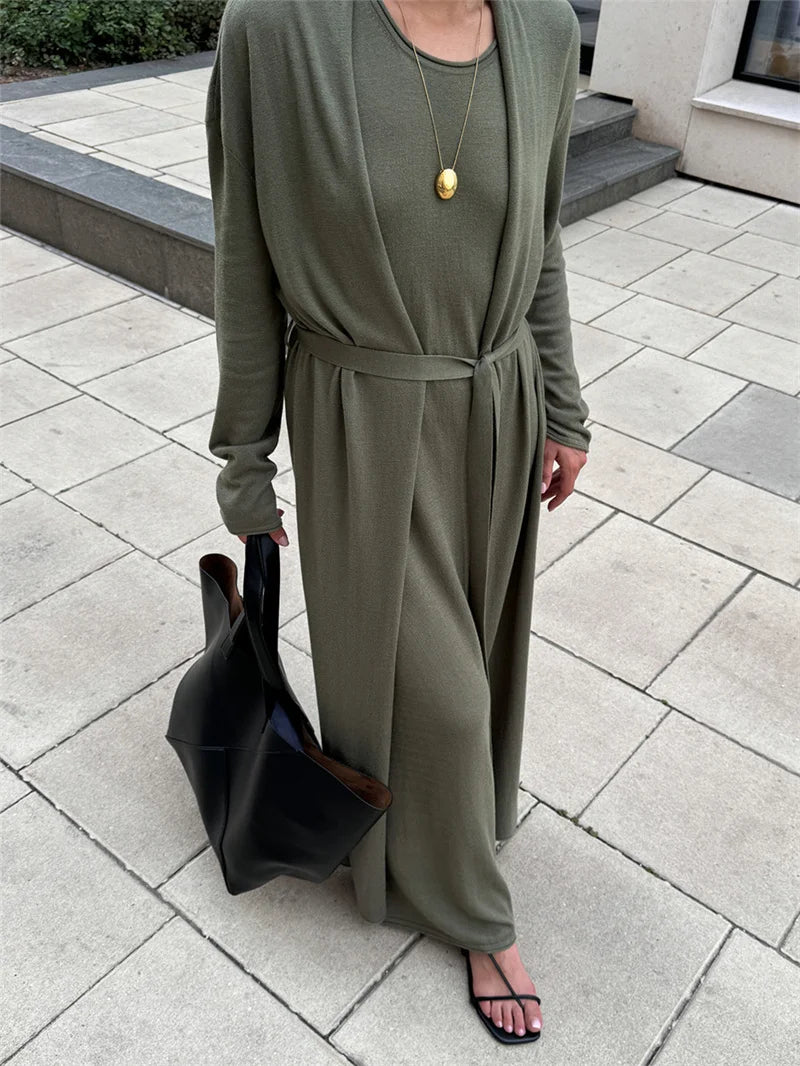 Person wearing a green long-sleeve dress with a belt, holding a black bag on a paved outdoor floor.