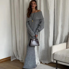 Woman wearing a long gray sweater holding a black handbag in a room with curtains and a sofa.