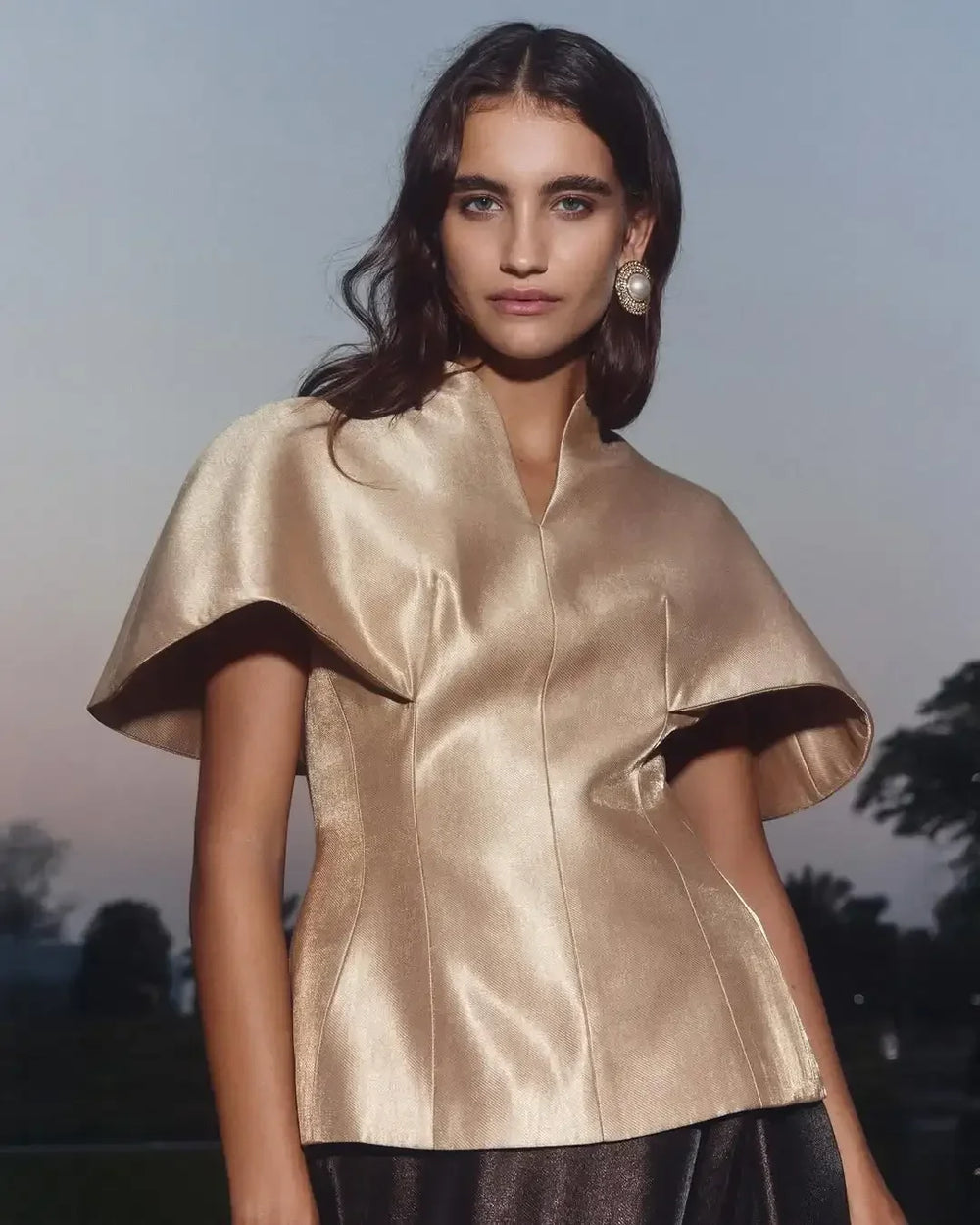 Woman wearing a gold satin top with a blurred outdoor background