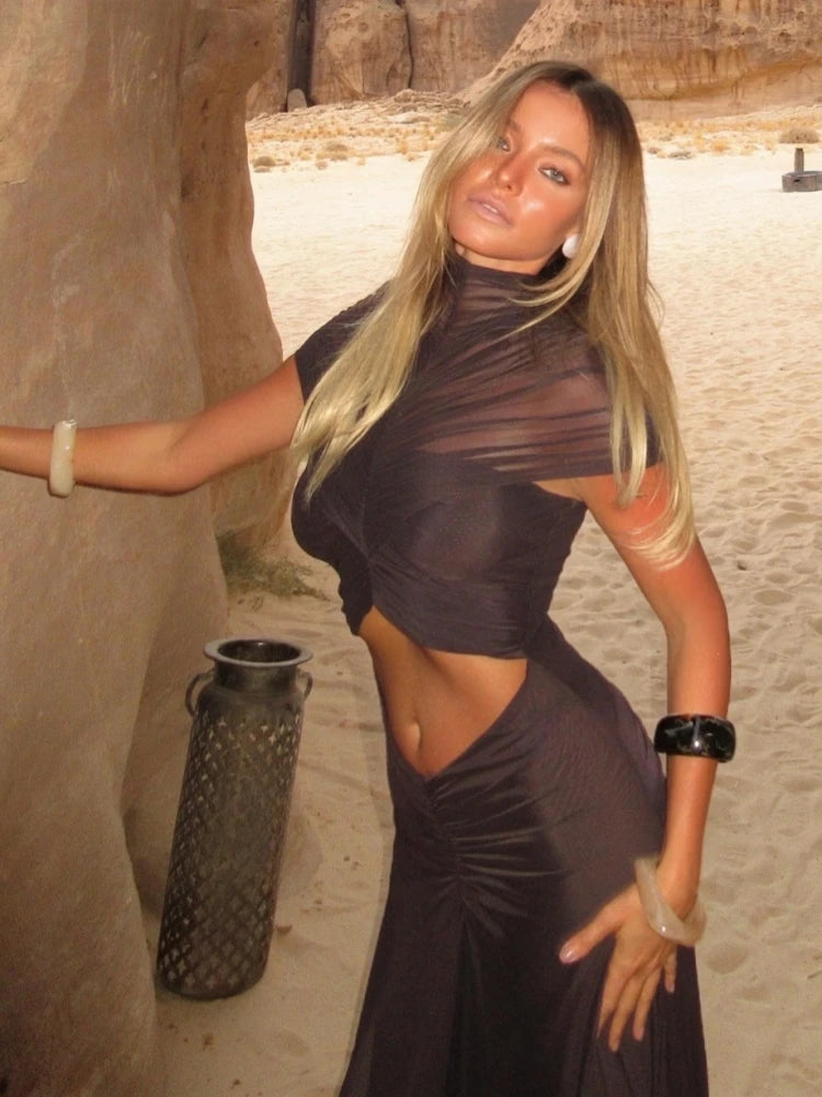 Woman in a dark dress standing in a desert-like setting with a textured wall.
