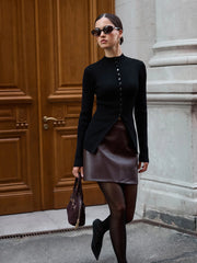 Woman in black top and brown leather skirt standing in front of a wooden door.