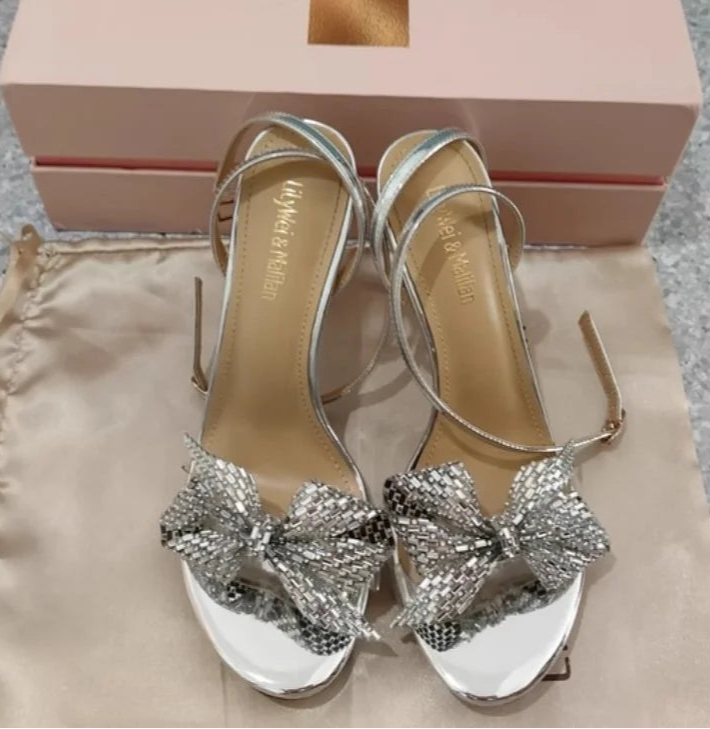 Silver high-heeled sandals with bow details on a beige surface, next to a pink shoe box.