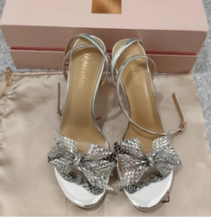 Silver high-heeled sandals with bow details on a beige surface, next to a pink shoe box.