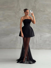 Woman in a black strapless dress with sheer overlay taking a selfie against a plain background