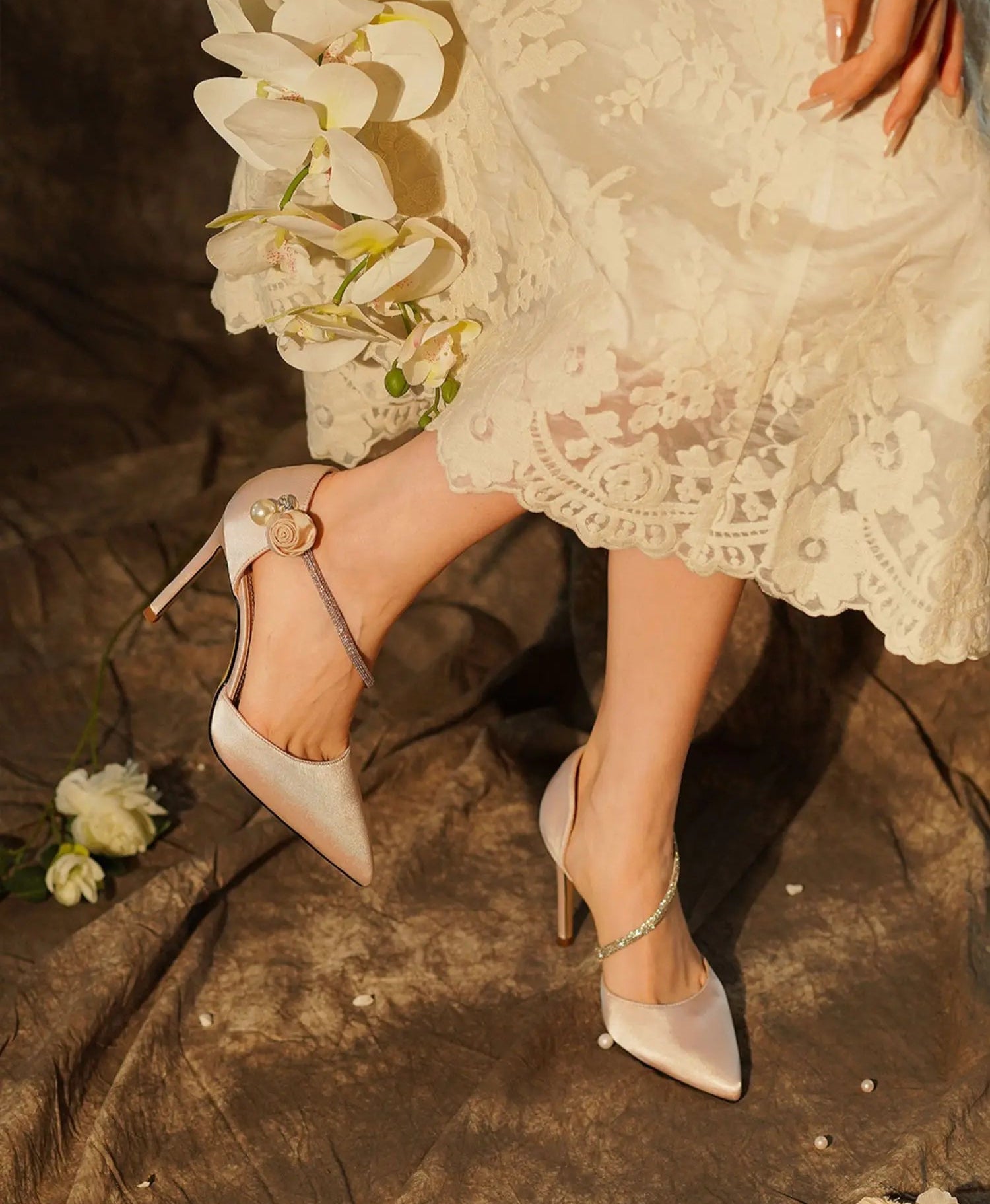 White high-heeled shoes with gold buckles worn by a person in a lace dress on a textured surface.