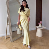 Woman in a yellow dress standing next to a mirror in a room with a table and vase.