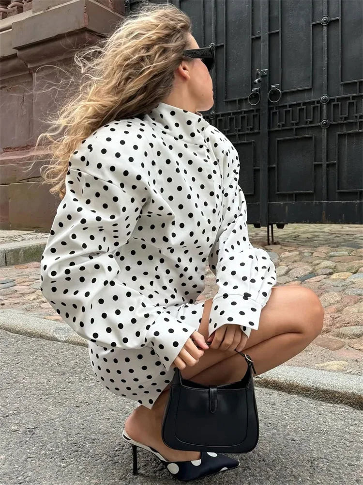 Woman in a polka dot blouse squatting on a street, holding a black handbag.