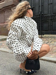 Woman in a polka dot blouse squatting on a street, holding a black handbag.